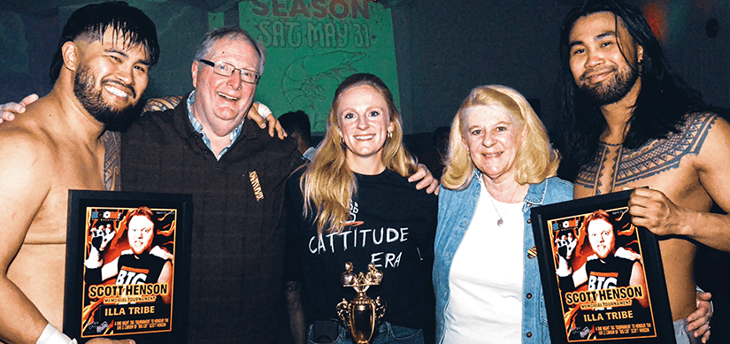 Two wrestlers holding commemorative photos of Scott Henson in his wrestling gear, standing on either side of the tournament trophy with Scott's sister and parents, smiling together.