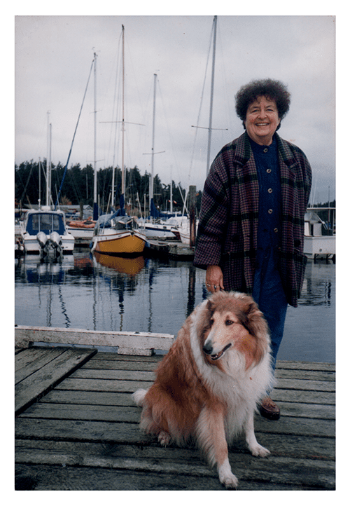 Forever Guardian Joan Hafeez standing on a marina dock on a cloudy day, with a big fluffy collie at her side.