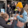 Woman wearing glasses gives a calm puppy a parvo vaccine being held by their guardian. The puppy is being held by a woman wearing a puffy teal jacket and yellow toque.