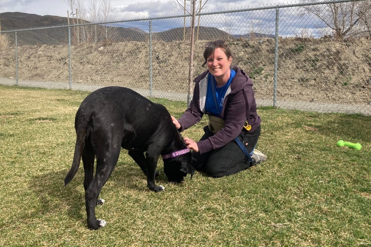 A picture of Rae smiling while petting a black dog against a fenced green field.
